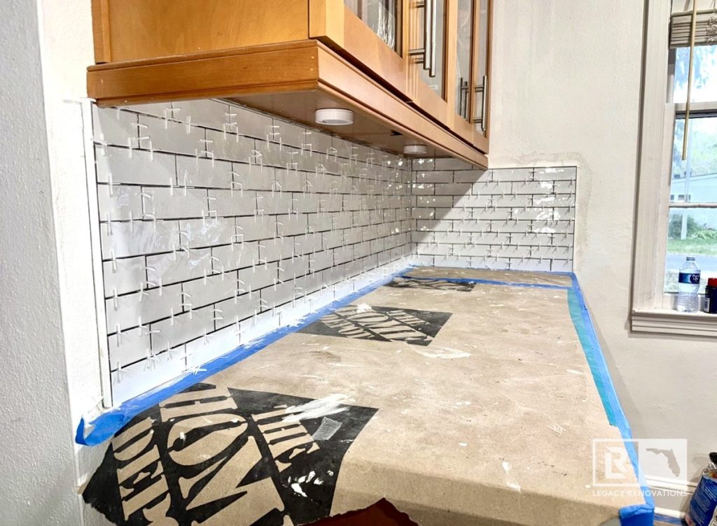 Kitchen remodel in Jacksonville, FL featuring a white subway tile backsplash being installed with tile spacers, natural wood cabinets, under-cabinet lighting, and protected countertops.