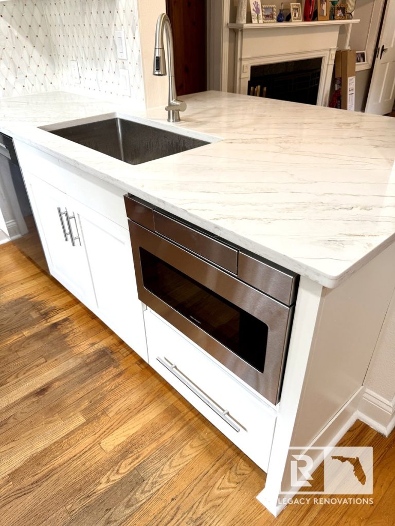 Kitchen remodel in Jacksonville, FL featuring a quartz peninsula, built-in stainless steel microwave drawer, farmhouse sink, pull-down faucet, white shaker-style cabinets, and hardwood flooring.