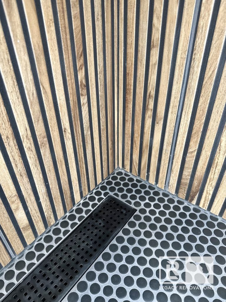Walk-in shower featuring a matte black linear drain, black penny round mosaic floor tile, and fluted wood-look wall tile in Jacksonville, FL.