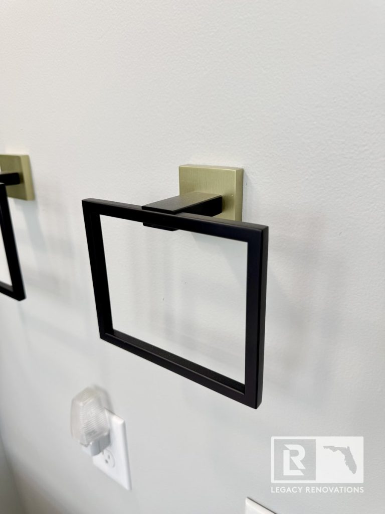 Close-up of a modern matte black rectangular towel ring with a brushed gold square bracket in a Jacksonville, FL bathroom renovation by Legacy Renovations.
