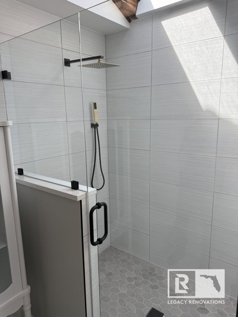 Modern walk-in shower with grey hexagon floor tiles, white textured wall tiles, matte black hardware, rainfall shower head, and natural light from a skylight, by Legacy Renovations in Jacksonville, FL.