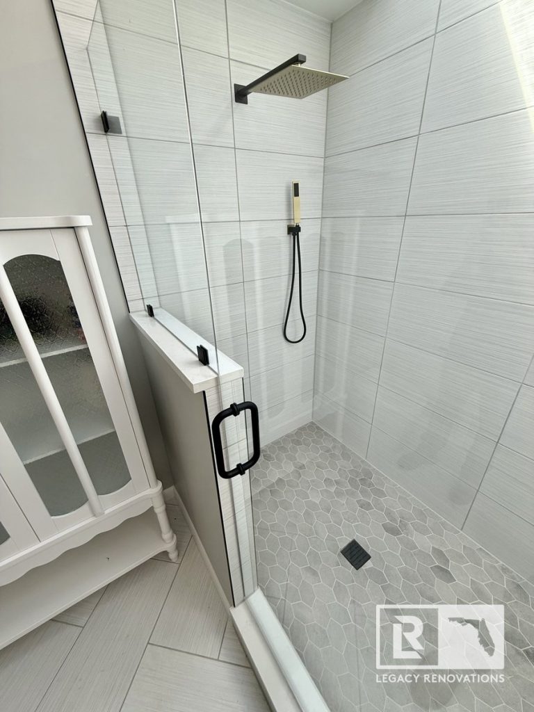 Modern walk-in shower with textured white wall tiles, grey hexagon floor tiles, and matte black fixtures in a Jacksonville, FL home remodel by Legacy Renovations.