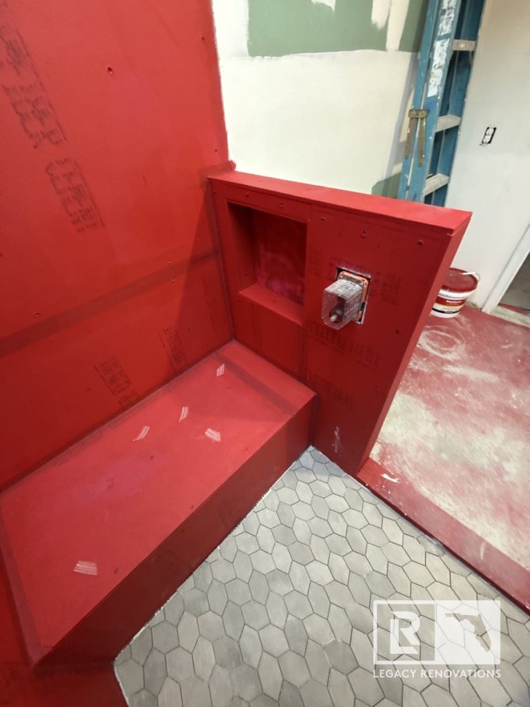 A walk-in shower renovation in progress featuring a grey hexagon tile floor alongside a built-in bench, pony wall, and niche coated in red waterproof membrane by Legacy Renovations in Jacksonville, FL.