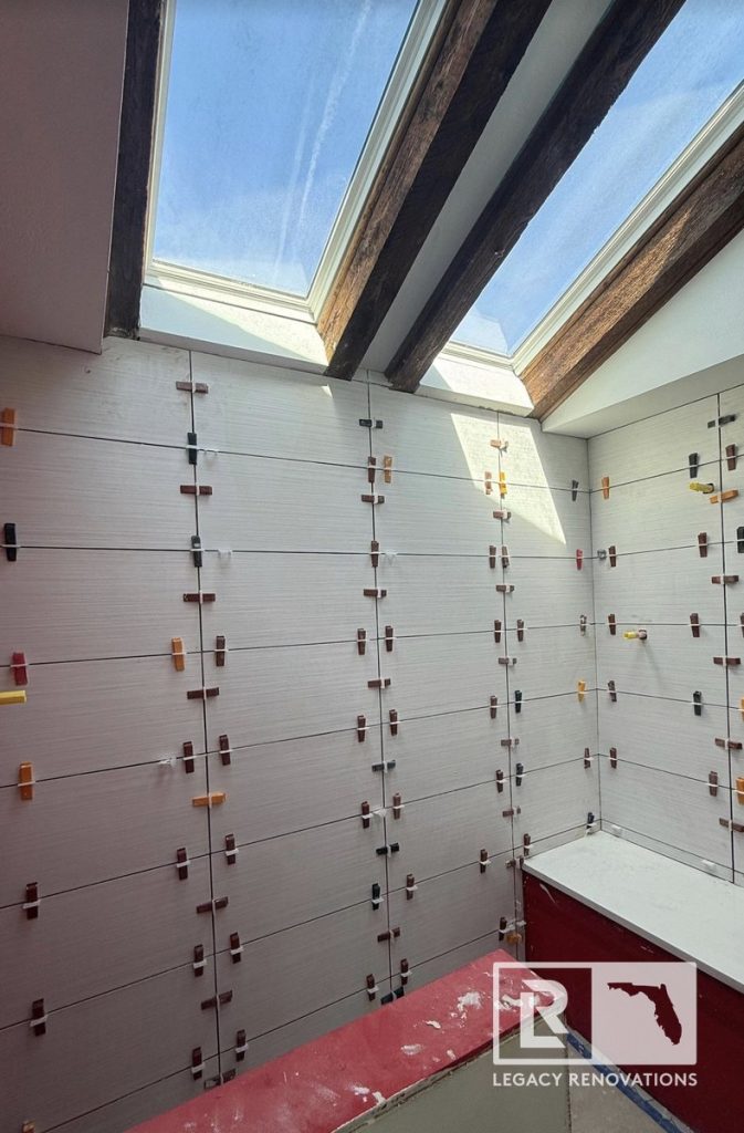 Bathroom remodel showing horizontal textured grey wall tiles with leveling spacers under twin skylights and rustic wood beams by Legacy Renovations in Jacksonville, FL.