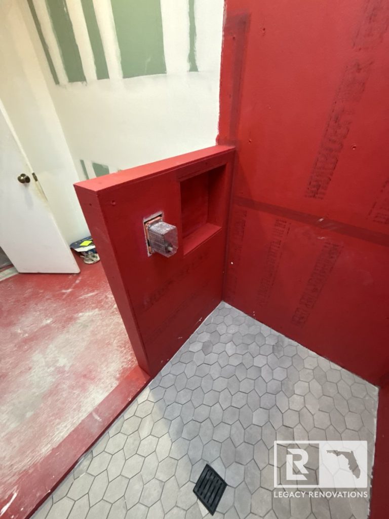A walk-in shower under construction with a grey hexagon tile floor, black square drain, rough-in plumbing valve, and red waterproof membrane on the framing by Legacy Renovations in Jacksonville, FL.