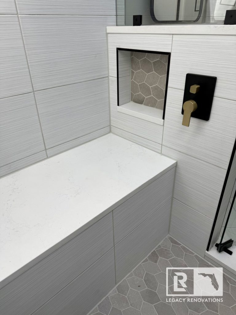 Built-in shower bench with a white quartz seat, textured grey wall tiles, a recessed hexagon-tiled niche, and modern gold shower controls in a Jacksonville, FL bathroom renovation.