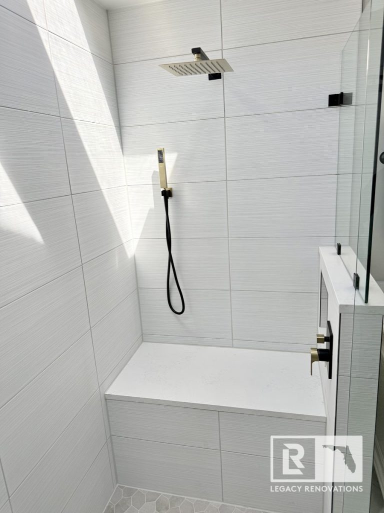 Walk-in shower featuring a white quartz bench, textured wall tiles, a square rainfall showerhead, and a handheld sprayer by Legacy Renovations in Jacksonville, FL.