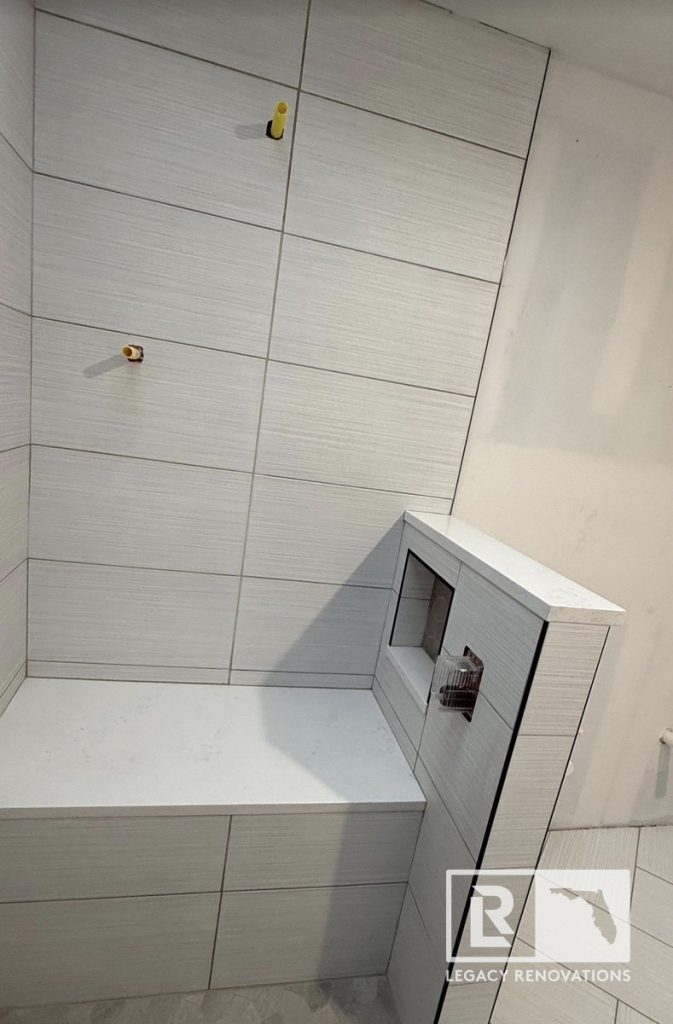 A walk-in shower during construction featuring textured grey wall tiles, a white stone bench seat, a built-in niche, and capped pony wall by Legacy Renovations in Jacksonville, FL.