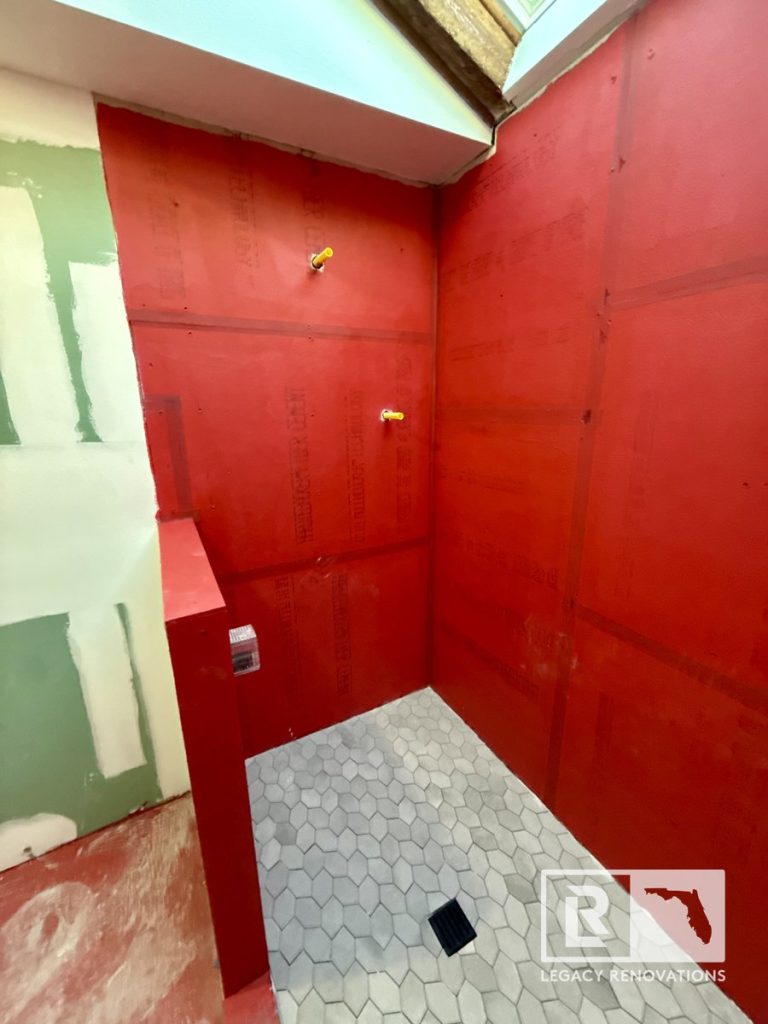 An in-progress bathroom renovation featuring a walk-in shower with grey hexagon floor tiles, a black square drain, red waterproofed backer boards, and plumbing cutouts by Legacy Renovations in Jacksonville, FL.