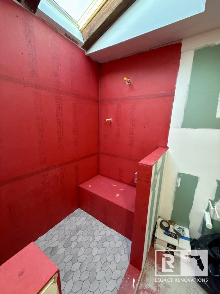An in-progress bathroom renovation featuring a walk-in shower with a built-in bench, bright red waterproofing membrane on the walls, grey hexagon tile floor, and an overhead skylight by Legacy Renovations in Jacksonville, FL.