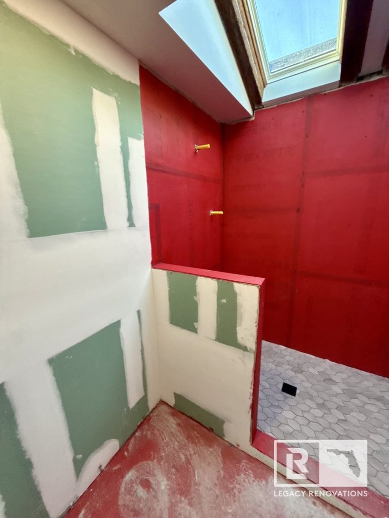 A bathroom remodel in progress showing a walk-in shower with bright red waterproofing membrane, grey hexagon floor tile, and unfinished green drywall panels by Legacy Renovations in Jacksonville, FL.