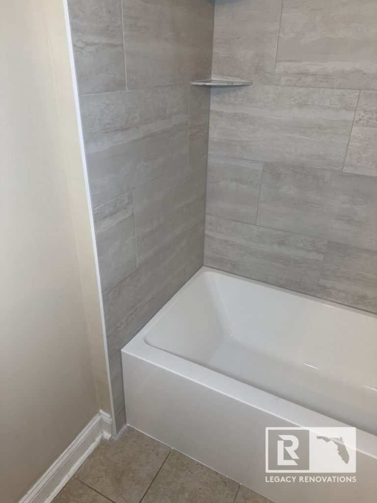 High-angle close-up of a glossy white acrylic alcove bathtub with light grey porcelain wall tile and a built-in corner shelf in a Jacksonville, FL bathroom.