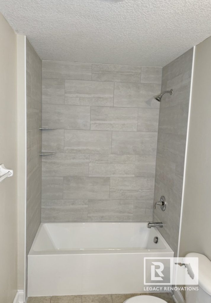 Modern bathroom featuring a white bathtub, large-format gray porcelain tile surround, marble corner shelves, and chrome fixtures in Jacksonville, FL.