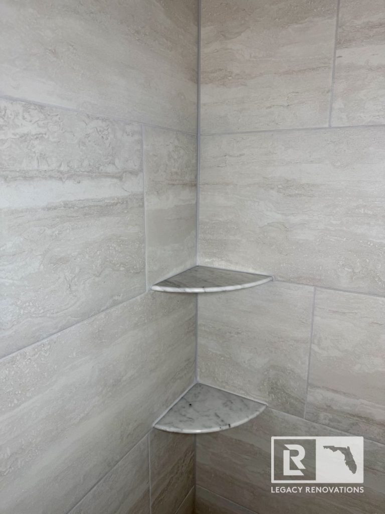Custom corner marble shower shelves installed with large-format gray porcelain tile during a bathroom remodel in Jacksonville, FL.