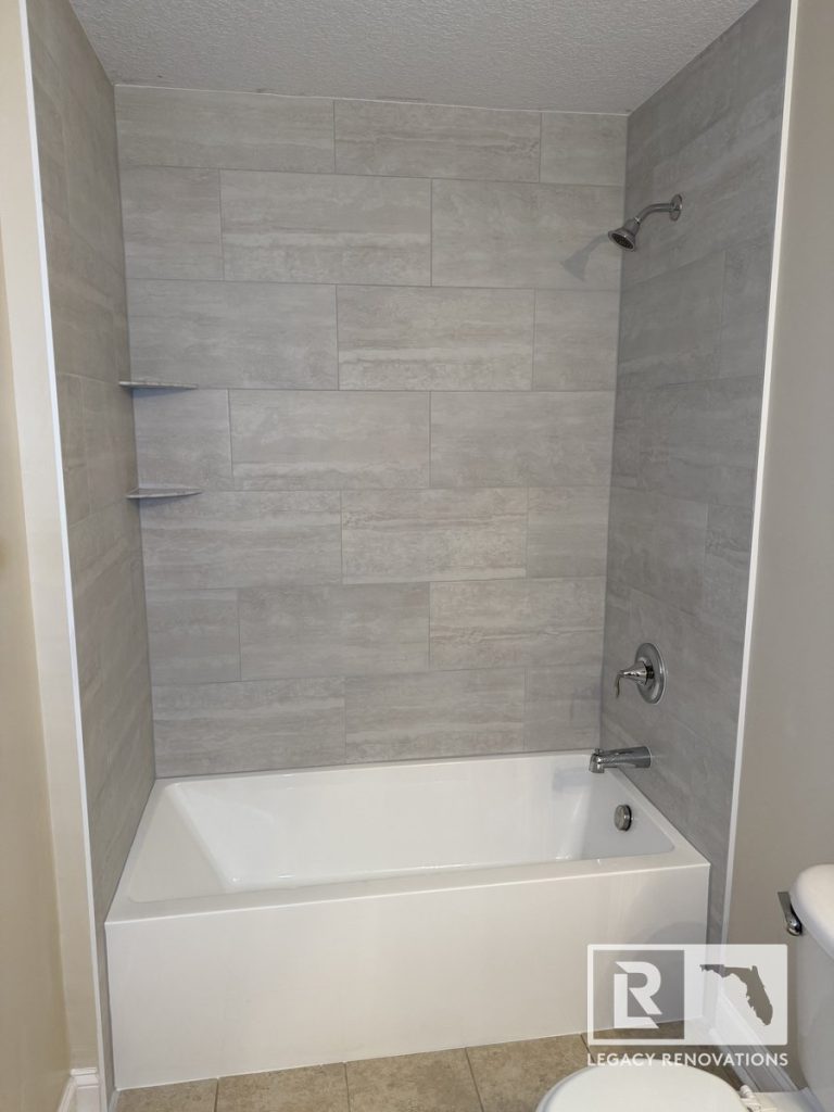 High-angle view of a clean white soaking bathtub with horizontal grey porcelain tile walls, chrome fixtures, and corner shelves in a Jacksonville, FL bathroom.