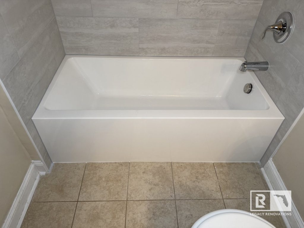 Straight-on view of a clean white alcove soaking tub, chrome fixtures, grey horizontal porcelain wall tile, and tan floor tile in a Jacksonville, FL bathroom.