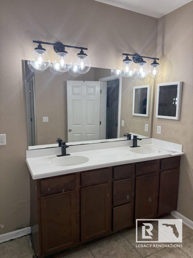 Before bathroom remodel in Jacksonville, Florida showing the original double-sink vanity, large wall mirror, builder-grade cabinets, quartz vanity top, and existing light fixtures before remodeling.