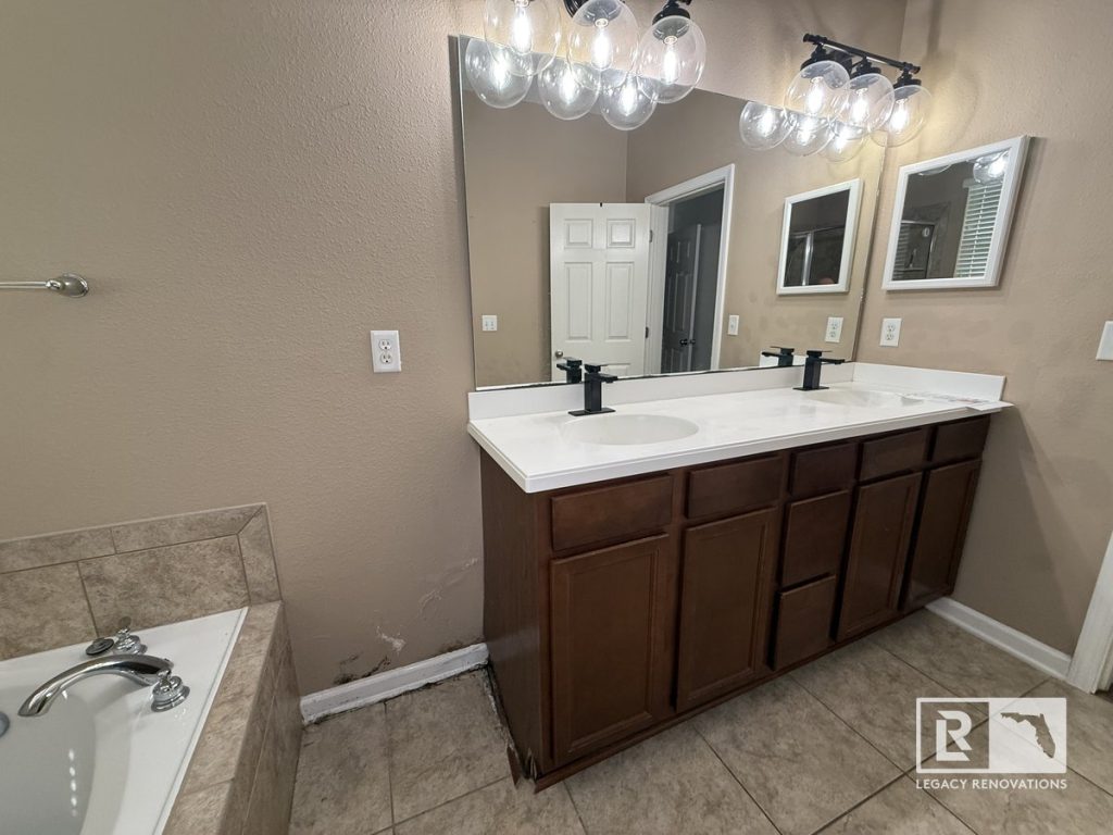 Before bathroom remodel in Jacksonville, Florida showing the original double vanity with dual sinks, builder-grade bathtub, tile flooring, and existing bathroom fixtures before renovation.
