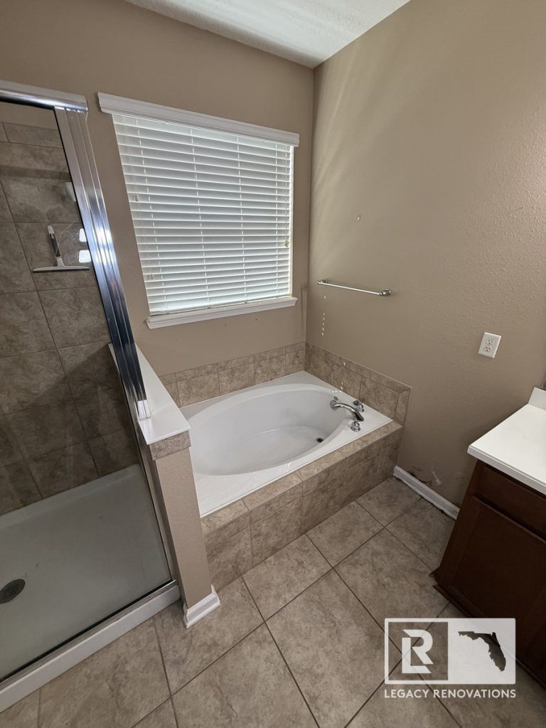 Original primary bathroom before renovation in Jacksonville, Florida showing a built-in soaking tub with ceramic tile surround, adjacent shower enclosure, window, and existing builder-grade finishes.