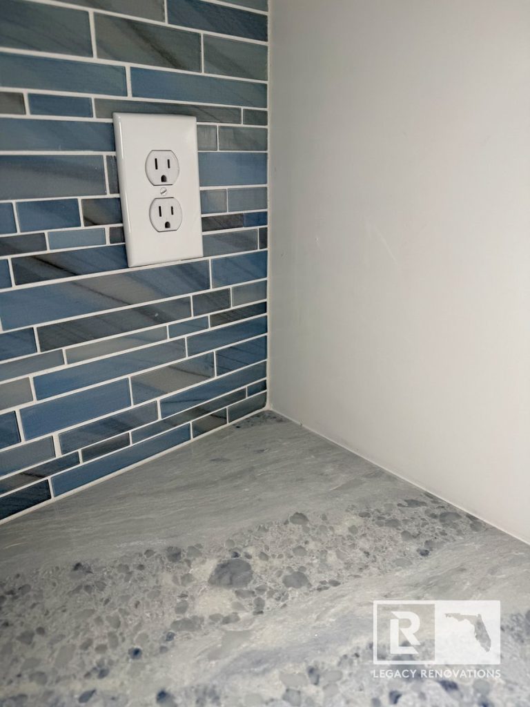 Close-up of a kitchen featuring a blue glass mosaic tile backsplash, gray-veined quartz countertop, clean corner finish, and integrated electrical outlet in a Jacksonville, FL kitchen remodel.