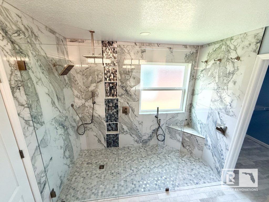 High-angle full view of a completed luxury walk-in shower with continuous marble-look porcelain tile, frameless glass enclosure, rain head, dual sprayers, and blue mosaic niche in Jacksonville, FL.