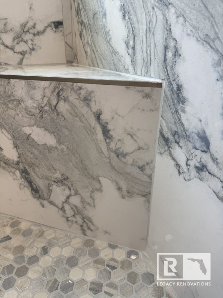Close-up of a custom shower bench with grey-veined marble porcelain tile and brass metal trim over mosaic tile floor in a Jacksonville, FL bathroom.