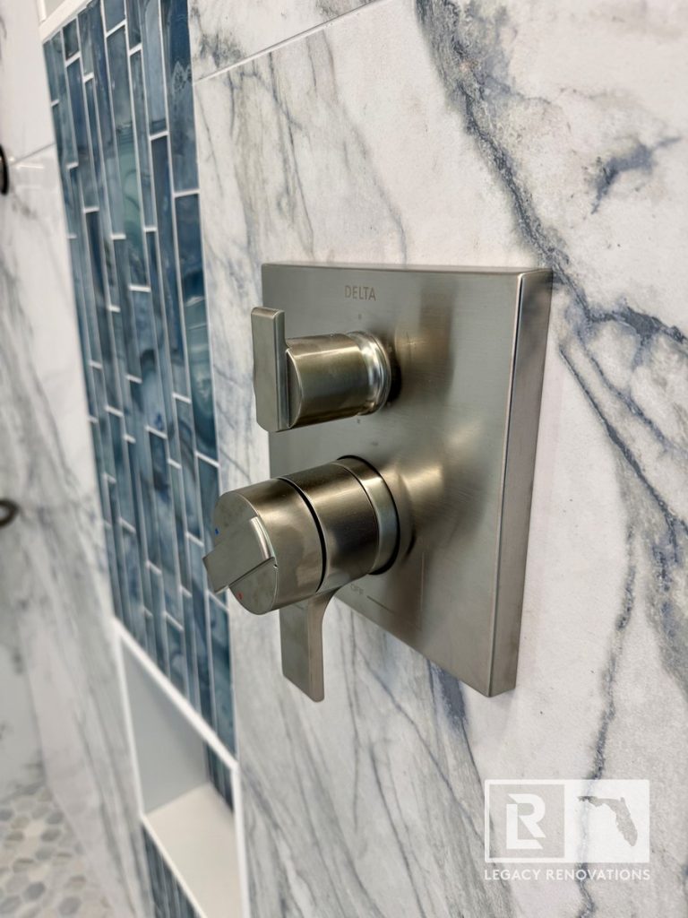Close-up of a square Delta brushed nickel dual-control shower valve trim on marble-look tile beside a blue glass mosaic accent strip in Jacksonville, FL.