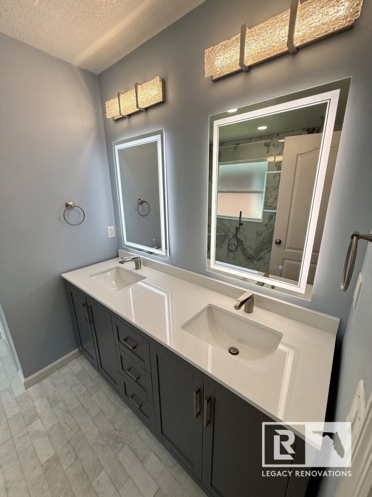 High-angle view of a dark grey double vanity with white quartz countertops, dual undermount sinks, illuminated LED mirrors, and modern light fixtures in a Jacksonville, FL bathroom.