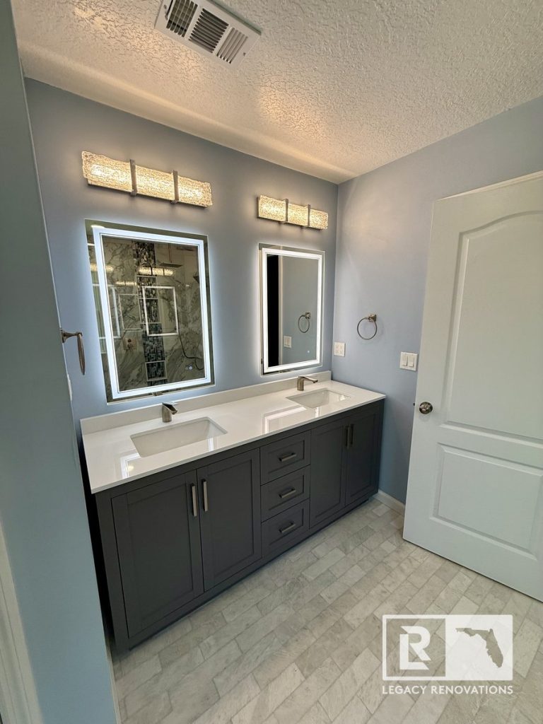 Modern bathroom remodel featuring a charcoal gray double vanity, quartz countertop, LED lighted mirrors, under-mount sinks, brushed nickel faucets, and stone-look tile flooring in a Jacksonville, Florida home.