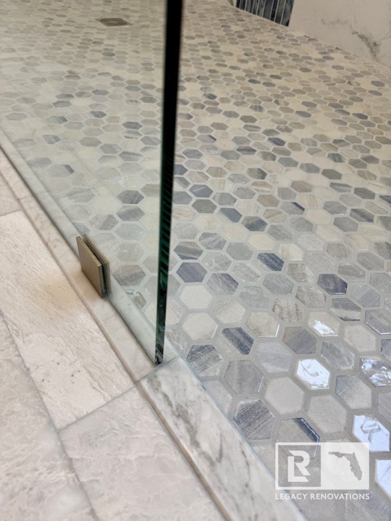 Close-up of a frameless glass shower panel anchored with a brushed bronze clip onto a marble threshold over hexagon floor tile in Jacksonville, FL.