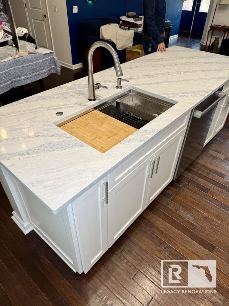 Kitchen remodel featuring a large island with a gray-veined quartz countertop, integrated stainless steel workstation prep sink, bamboo cutting board, pull-down faucet, built-in storage cabinets, and hardwood flooring in Jacksonville, FL.