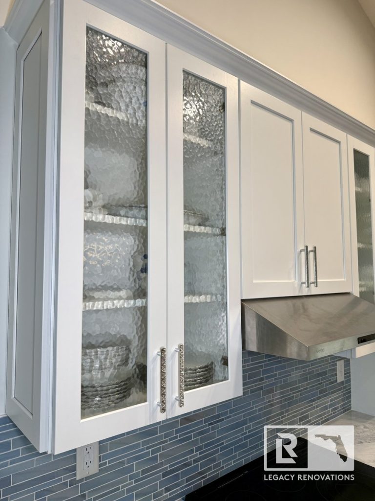 Kitchen featuring glass-front display cabinets with textured glass panels, brushed nickel handles, stainless steel range hood, and blue glass tile backsplash in a Jacksonville, FL home renovation.