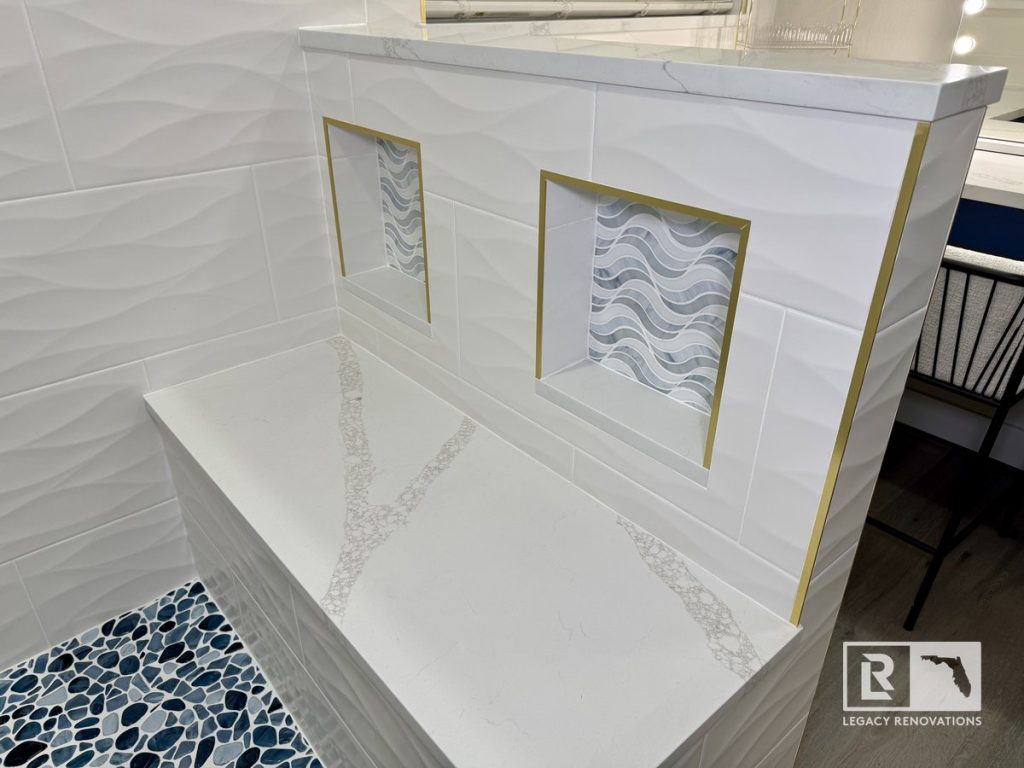 Custom bathroom remodel in Jacksonville FL featuring a quartz shower bench, decorative shower niches with gold trim, textured white wall tile, and blue pebble shower floor in a modern luxury bathroom.
