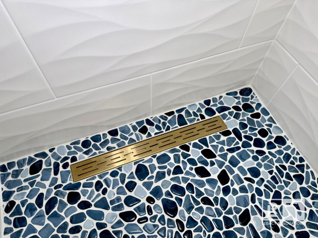 Custom walk-in shower floor with blue pebble mosaic tile, brushed brass linear drain, and textured white wall tile in a Jacksonville, Florida bathroom remodel.
