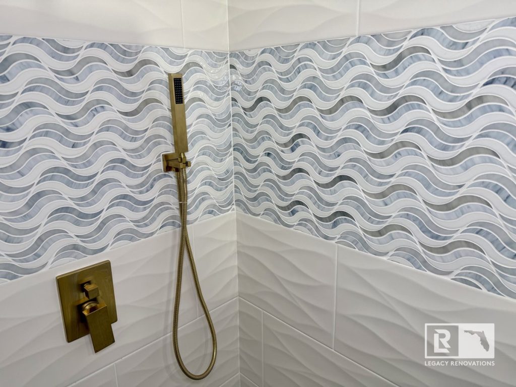 Walk-in shower with a blue and gray wave mosaic accent tile, textured white wall tile, and brushed brass fixtures in a Jacksonville, Florida bathroom remodel.
