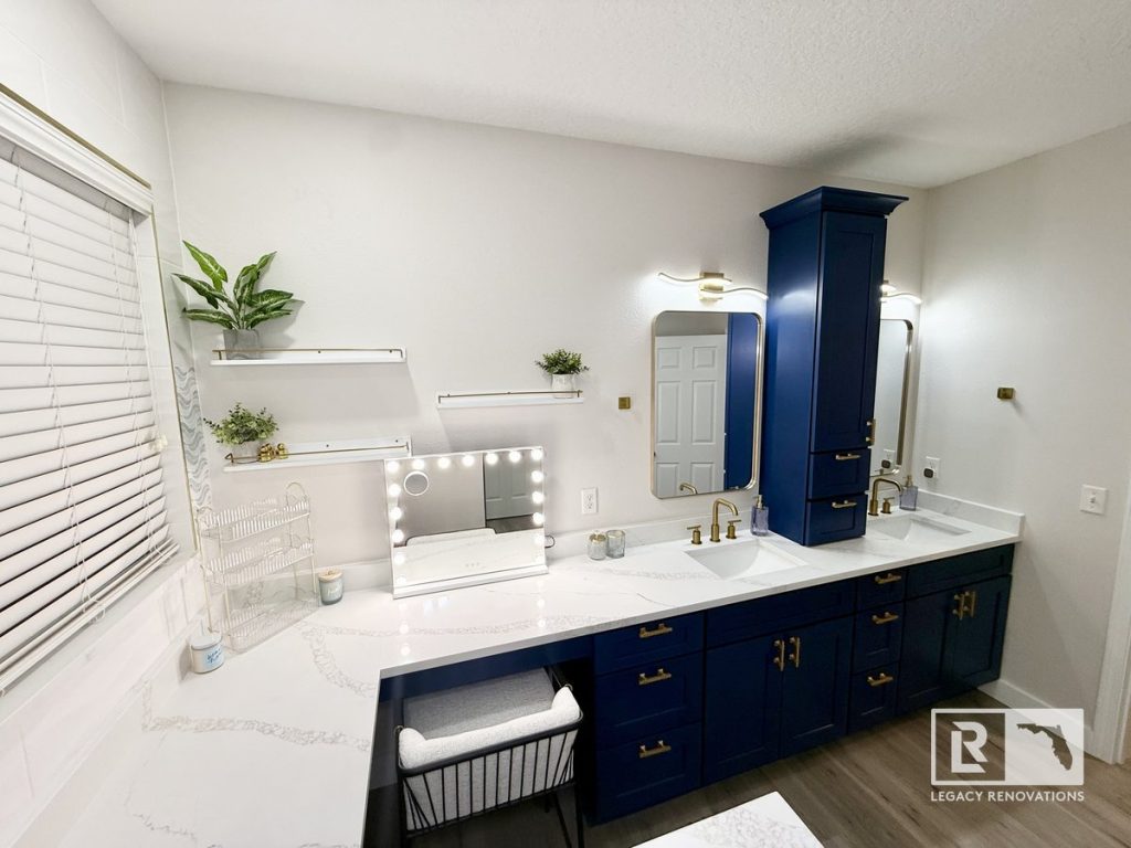 Luxury bathroom remodel in Jacksonville, Florida featuring a custom navy blue double vanity, white quartz countertops, brushed brass fixtures, a built-in makeup station, and a full-height storage cabinet.