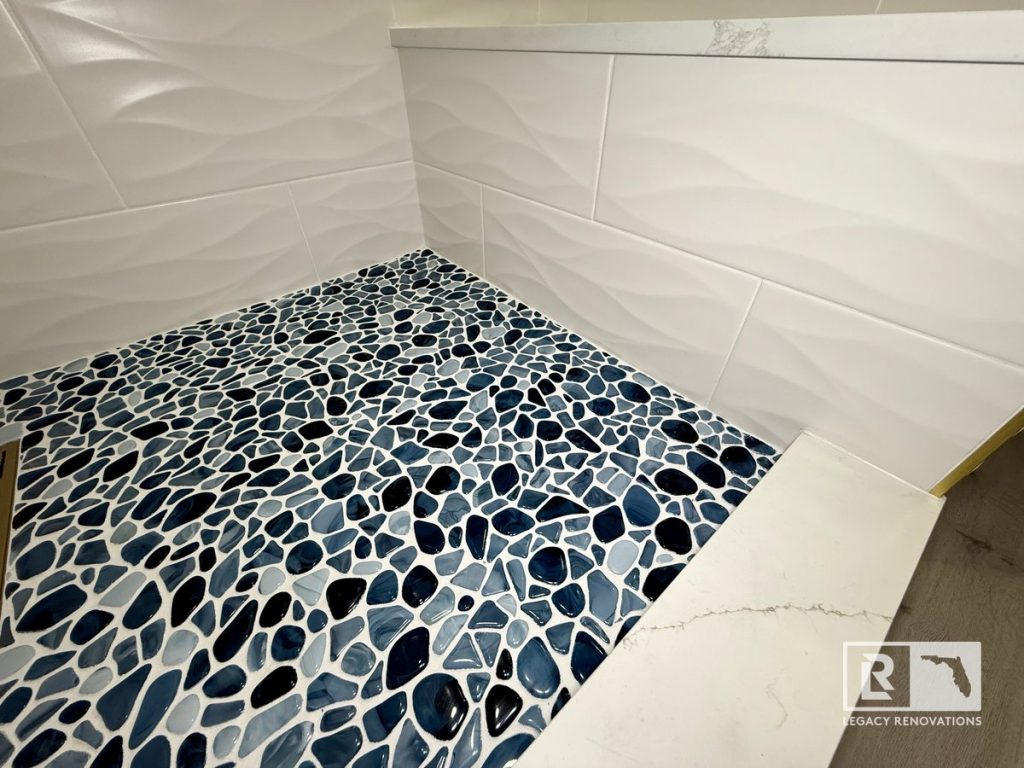 Blue pebble shower floor tile in a Jacksonville FL bathroom remodel featuring textured white wall tile, quartz shower bench, and custom walk-in shower design.