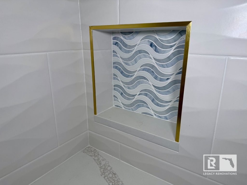 Custom shower niche in Jacksonville FL bathroom remodel featuring blue wave mosaic tile, brushed gold trim, textured white wall tile, and luxury custom shower design.