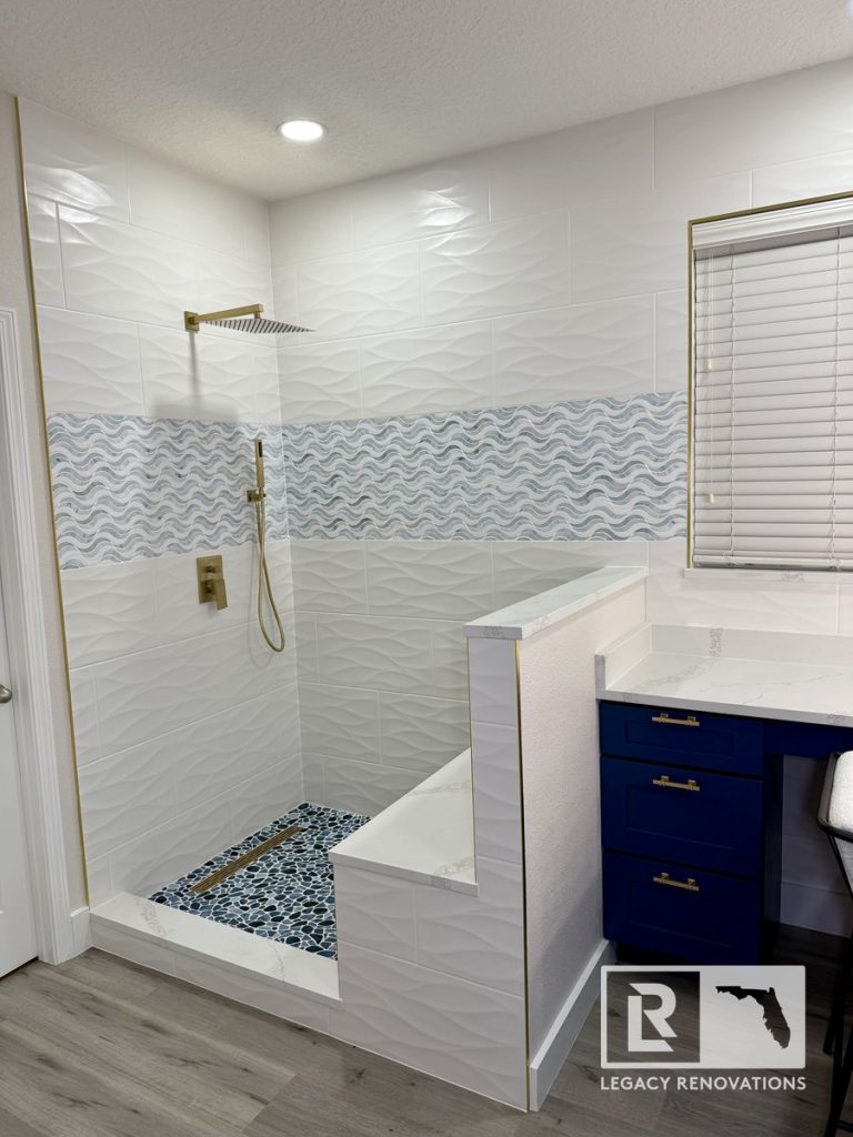 Walk-in shower remodel in Jacksonville, Florida featuring textured white wall tile, wave accent tile, brushed gold shower fixtures, pebble shower floor, and modern half-wall design.