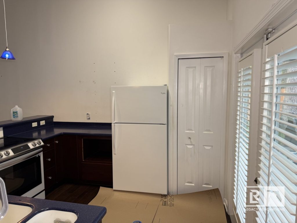 Before kitchen remodel showing dark wood cabinets, blue laminate countertops, white refrigerator, tall ceiling, pantry door, and large windows in a Jacksonville home.
