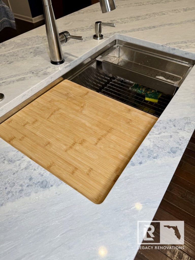 Close-up of a stainless steel kitchen workstation sink with a bamboo cutting board, drying rack, prep colander, pull-down faucet, soap dispenser, and gray-veined quartz countertop in Jacksonville, FL.