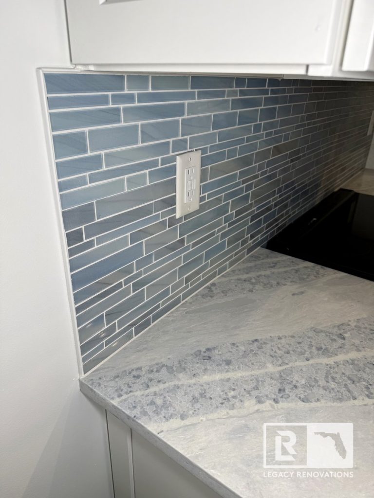 Kitchen detail featuring a gray-veined quartz countertop, blue linear glass tile backsplash, under-cabinet installation, and modern wall outlet in a Jacksonville, FL kitchen remodel.