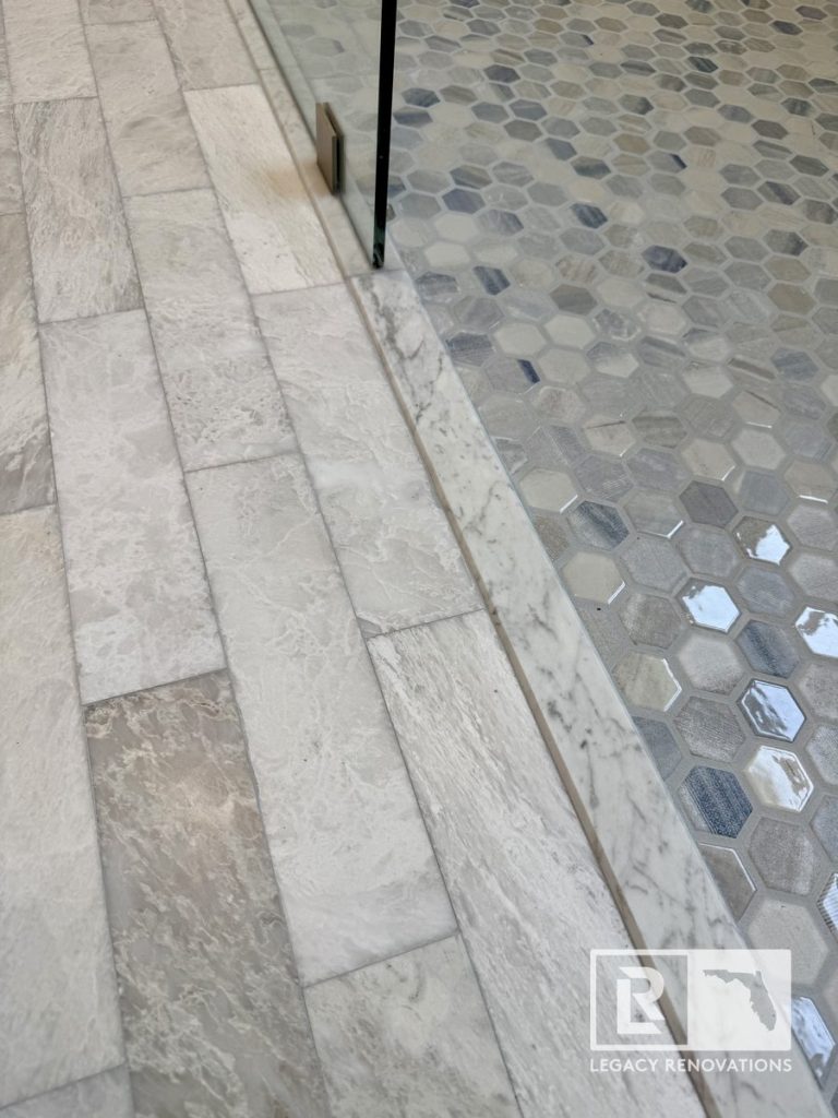 Close-up of a bathroom shower entrance featuring a marble threshold, frameless glass panel, stone-look porcelain floor tile, and gray hexagon mosaic shower floor in a Jacksonville, Florida home.