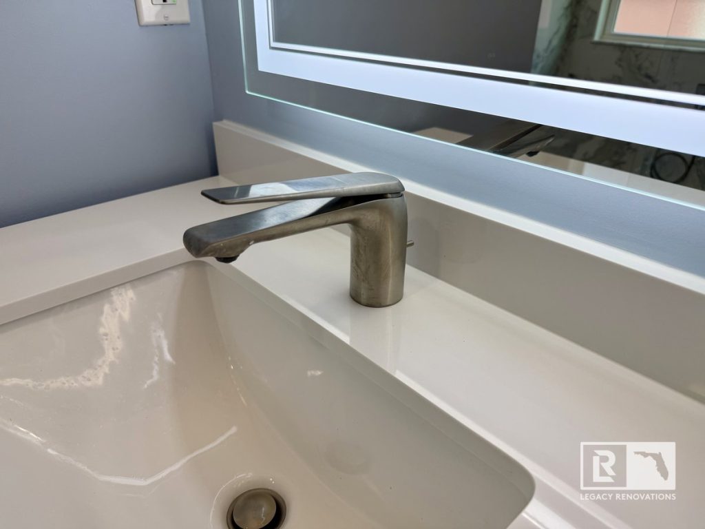 Close-up of a contemporary brushed nickel single-handle faucet, white quartz countertop, under-mount sink, and back-lit LED mirror in a Jacksonville, FL bathroom.