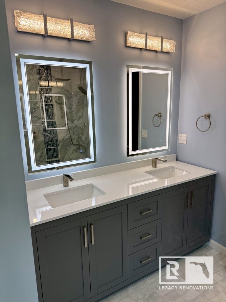 Modern bathroom with a charcoal gray double vanity, white quartz countertop, LED backlit mirrors, brushed nickel faucets, and a marble-look walk-in shower in Jacksonville, Florida.