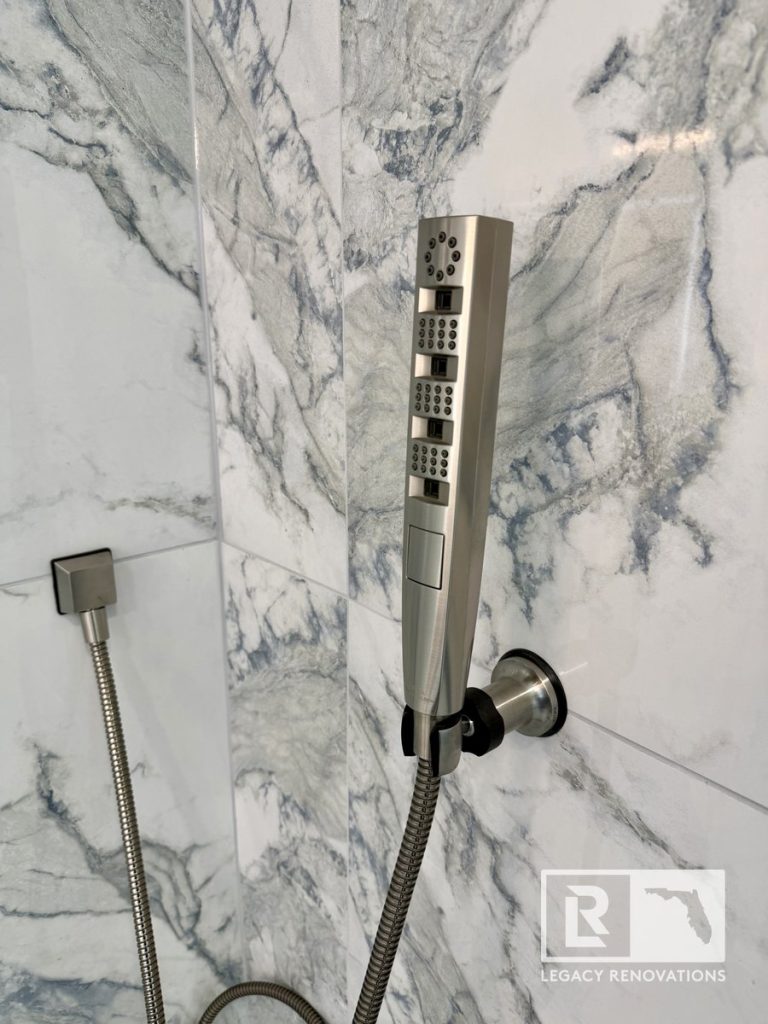 Close-up of a contemporary brushed nickel handheld shower wand mounted on grey and blue veined marble-look tile in a Jacksonville, FL bathroom.