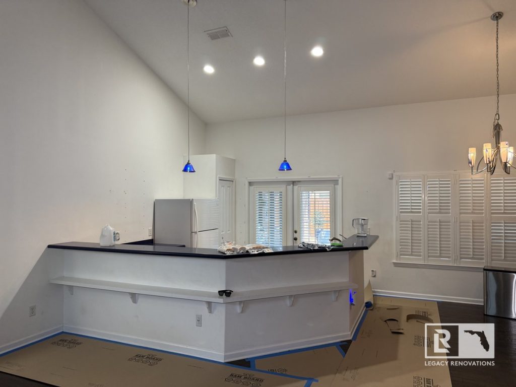 Wide view of an open-concept kitchen before renovation with a raised breakfast bar, high ceilings, pendant lighting, white refrigerator, and adjoining dining area in a Jacksonville, Florida home.