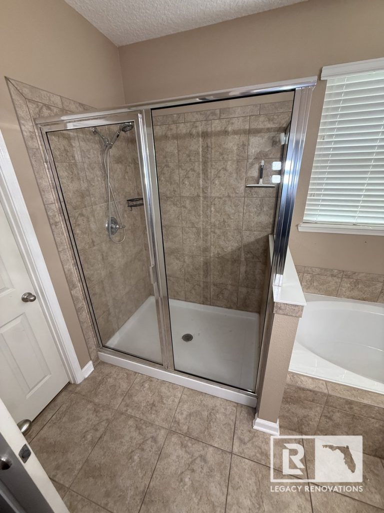 Original bathroom shower in Jacksonville, Florida with a framed sliding glass enclosure, ceramic tile walls, built-in corner shelf, and adjacent soaking tub before renovation.