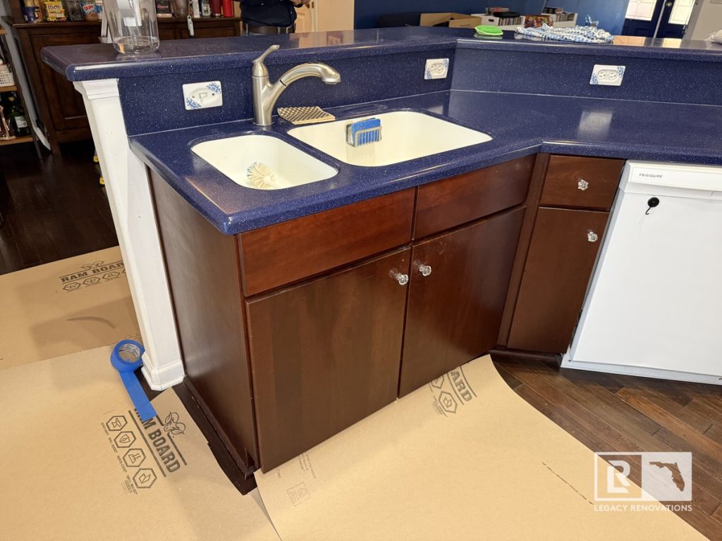 Original kitchen with dark wood base cabinets, blue countertops, double-basin sink, peninsula, and dishwasher before renovation in a Jacksonville, Florida home.