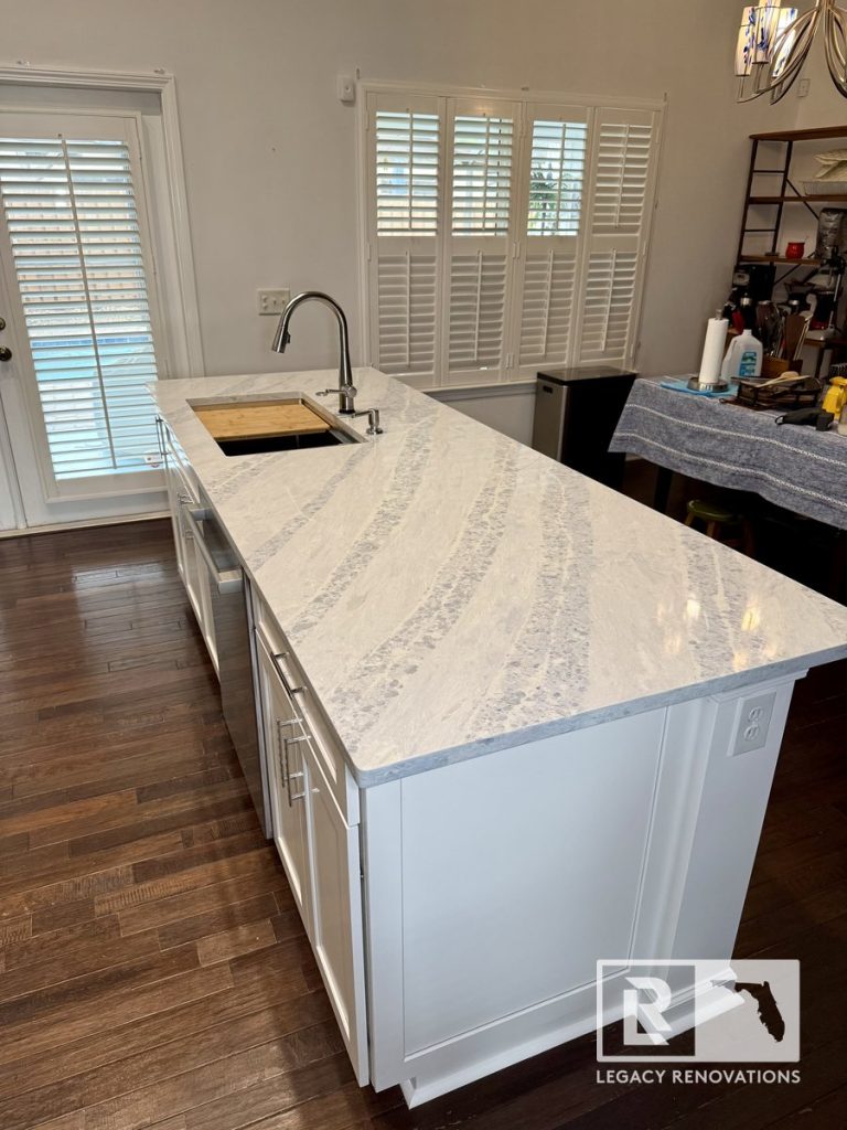 Contemporary kitchen featuring an oversized quartz island with gray veining, stainless steel workstation sink, pull-down faucet, hardwood floors, and plantation shutters in Jacksonville, FL.